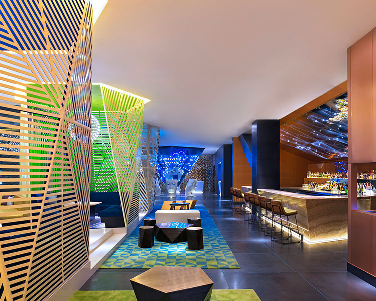 A modern, stylish bar interior featuring geometric patterned screens, a mix of green and blue accents, sleek high-top tables with stools, and a well-lit counter. The space combines contemporary design elements with ambient lighting and a futuristic ambiance.