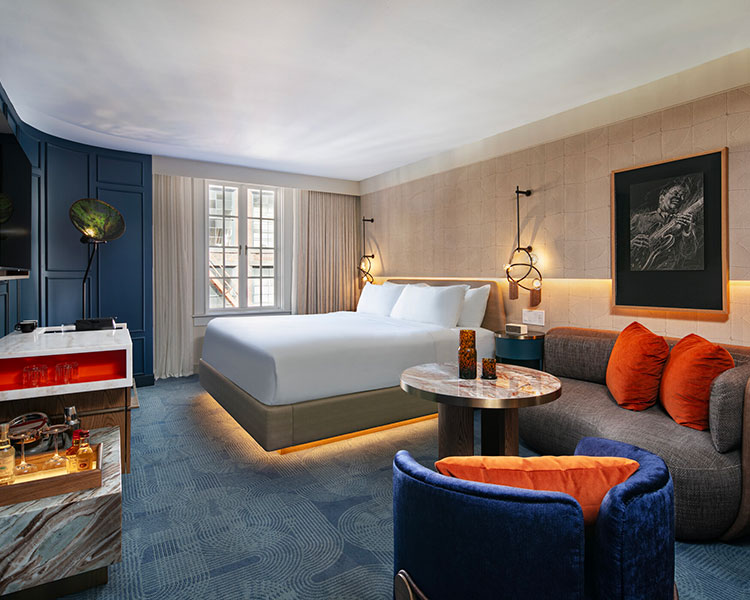 A modern hotel room featuring a large, neatly made bed with white linens. The room includes a blue armchair, a small round table, a grey sofa with orange cushions, and a well-lit desk. The decor features warm lighting, a wall painting, and a large window with sheer curtains.