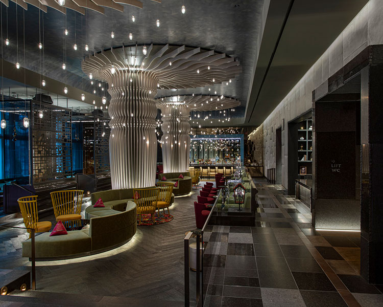 A stylish, modern hotel lobby featuring unique, wavy ceiling structures with pendant lights hanging overhead. The space has cozy seating areas with green sofas, yellow chairs, and decorative cushions. The sleek design includes a checkered floor and a bar area in the background.