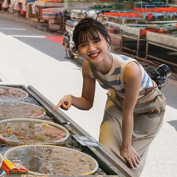 A woman smiles and points at seafood displayed in round baskets filled with water. She wears a striped tank top and has a camera slung over her shoulder. The scene appears to be at an outdoor seafood market.