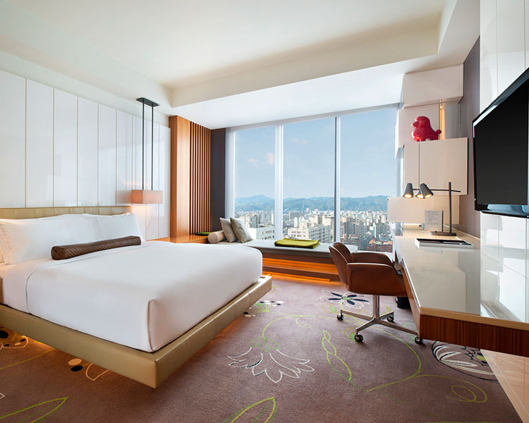 A modern hotel room with a large bed and white bedding, a wall-mounted TV, a desk and chair, and a window offering a city view with mountains in the background. The room has a minimalist design with neutral tones and a few decorative accents.