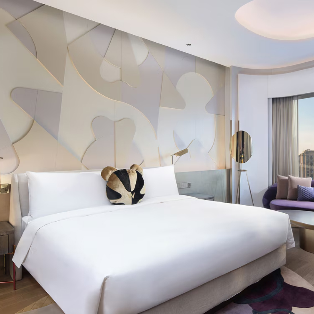 A modern hotel room with a large bed decorated with white linens and a black-and-gold heart-shaped pillow. The room features abstract wall art, a large floor lamp, a purple armchair with pillows, and a large window with a view of a cityscape.