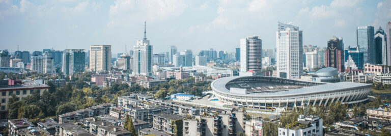 A panoramic view of an urban cityscape featuring a mix of modern high-rise buildings and older structures. In the foreground, a large stadium with an oval roof stands prominently. The sky is partly cloudy, casting a serene light over the city.