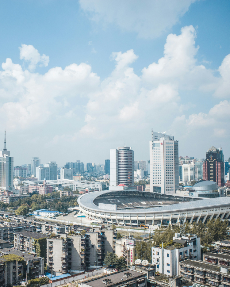 A panoramic view of a modern cityscape featuring high-rise buildings under a blue sky with scattered clouds. In the foreground, a large, elliptical stadium stands out among shorter, older buildings. The city extends into the distance, showcasing urban density.
