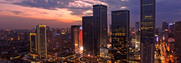 A panoramic view of a modern cityscape at dusk with tall skyscrapers illuminated by lights. The sky is painted with hues of orange and purple as the sun sets, casting a soft glow over the buildings and the bustling streets below.