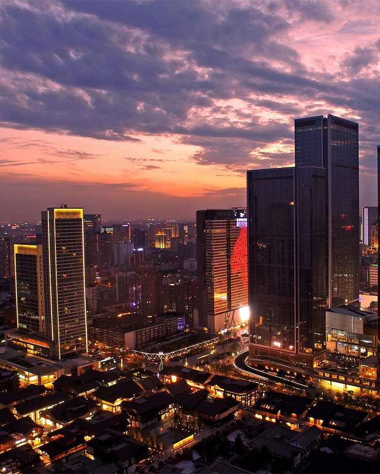 A city skyline at dusk featuring tall, illuminated buildings under a dramatic, colorful sky with streaks of orange and purple clouds. The city lights glow, reflecting off the buildings, creating a vibrant urban landscape.