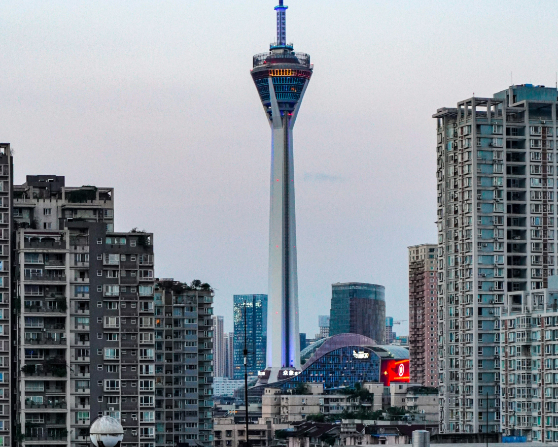 The image shows a skyline with a prominent tower featuring an observation deck in the middle, flanked by two tall, densely packed residential buildings. More high-rise buildings and a few illuminated signs are visible in the background, under a clear sky.
