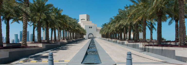 A wide walkway lined with tall palm trees leads to a modern, white building with geometric architecture. The path features a long, narrow fountain running down the center. The sky is clear and blue, creating a striking contrast with the structure and surroundings.