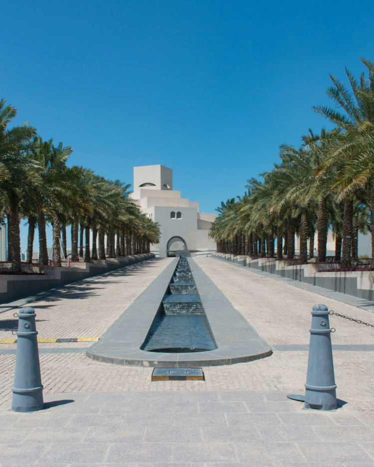 A long, symmetrical pathway lined with palm trees on both sides leads to a modern, white building under a clear, blue sky. A narrow, rectangular reflecting pool runs through the center of the pathway, bordered by clean geometric lines.