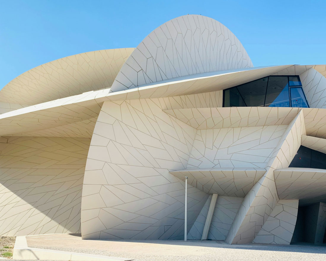 A modern architectural building with overlapping, curved panels and angular shapes resembling a desert rose. The facade is light-colored and textured, with a clear blue sky in the background. The design includes various geometric forms and large windows.