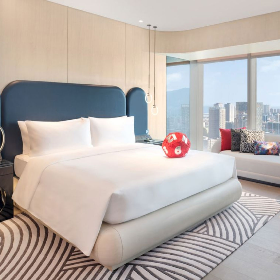 A modern hotel room with a large bed featuring white linens and a blue headboard. There is a red 