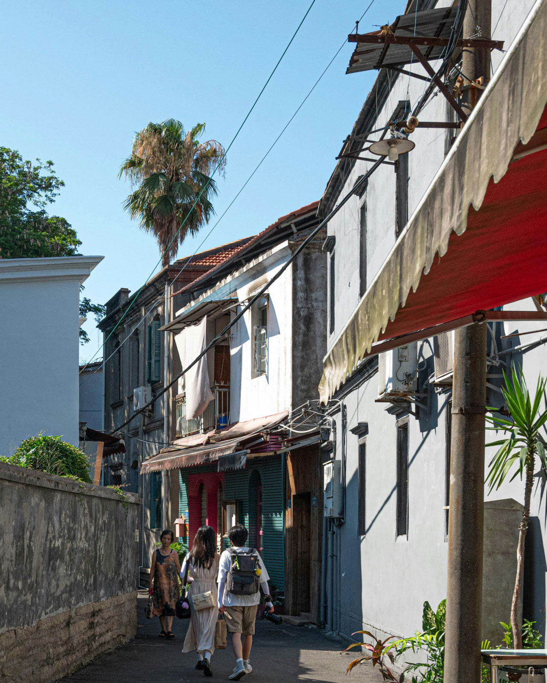 A narrow street with old, multi-story buildings under a bright blue sky. People walk along the path, with some greenery visible at the sides and a palm tree in the distance. The buildings have a mix of red and green doors, and an awning hangs from one building.