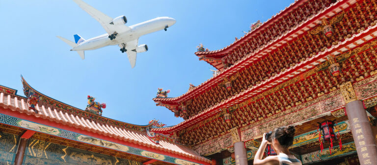 A large commercial airplane flies overhead against a clear blue sky. Below, a person is taking a photo of a traditional Asian temple with ornate, colorful architecture and detailed roof designs.