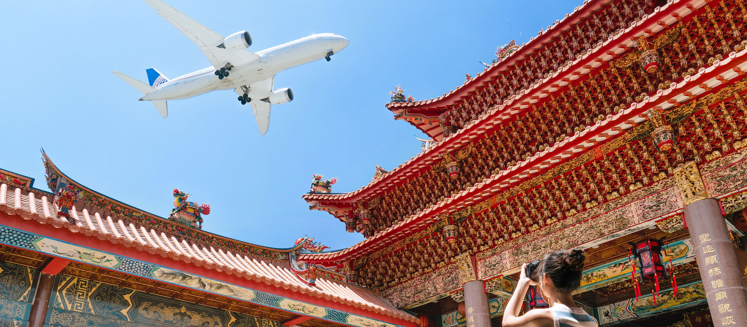 Une personne prend une photo d'un temple asiatique traditionnel richement décoré avec des touches de rouge et d'or. Au-dessus, un avion blanc vole au-dessus d'eux dans un ciel bleu clair.