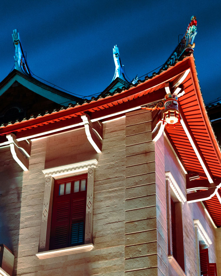 An ornate building shows traditional Asian architectural elements with curved rooflines and intricate detailing. Illuminated with soft lighting against a twilight sky, the structure's red and beige hues stand out, emphasizing its cultural and historic charm.