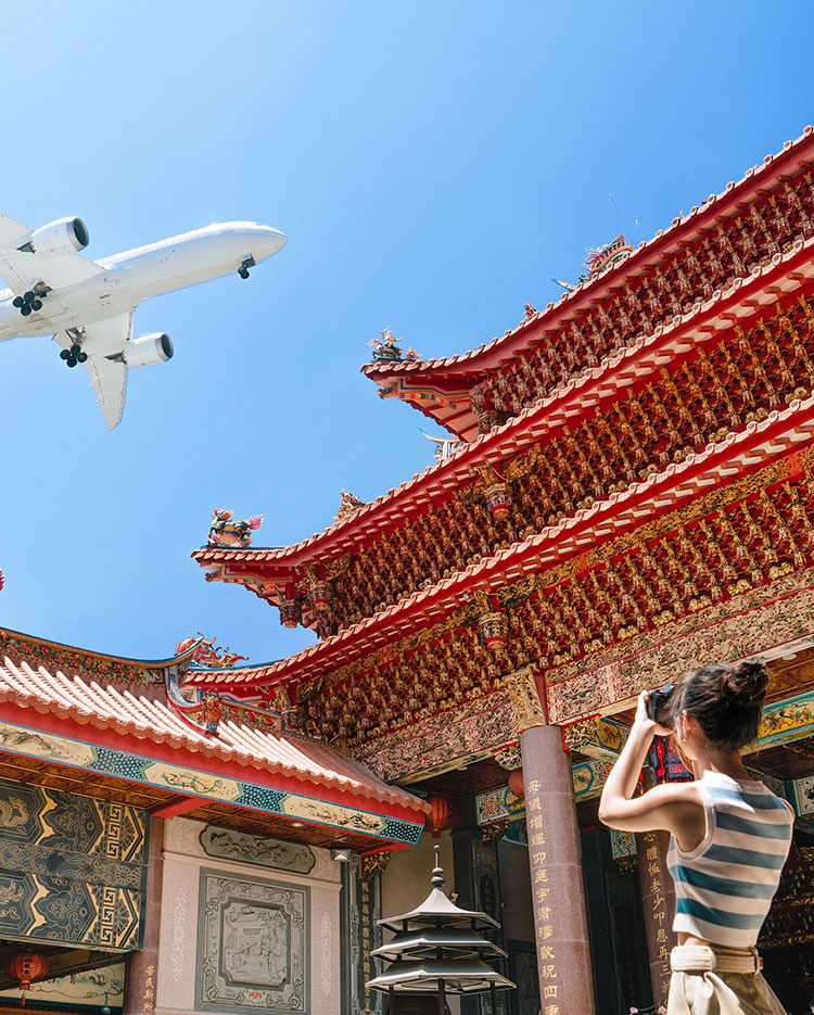 Eine Person mit einem Fernglas blickt zu einem großen, detailreichen traditionellen asiatischen Tempel hinauf. Ein Flugzeug fliegt über den klaren blauen Himmel hinweg und erzeugt einen Kontrast zwischen moderner Luftfahrt und antiker Architektur.