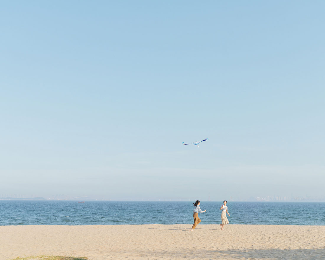 澄んだ青い空と穏やかな海を背景に、砂浜で凧揚げをしている2人。凧は青と白で、彼らの上空に舞い上がっています。穏やかで楽しい光景です。.