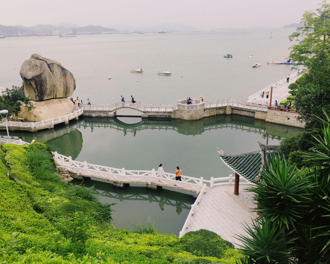 A scenic view of a coastal park featuring a large rock formation, a pond with white bridges, lush greenery, and a panoramic view of the calm sea with boats in the distance. People are walking along the pathways and enjoying the serene atmosphere.