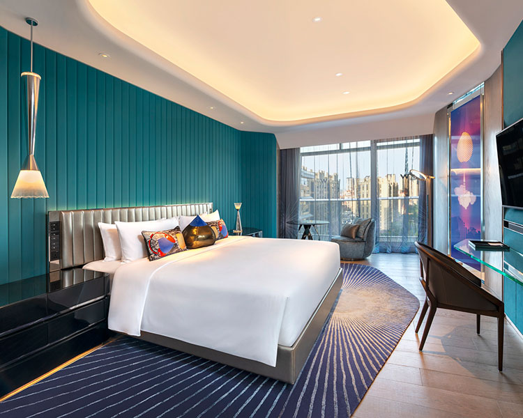 A modern hotel room featuring a large king-sized bed with white linens and colorful pillows. The room has teal walls, wooden floors, and floor-to-ceiling windows with city views. There is a sleek desk, lounge chair, and unique lighting fixtures.