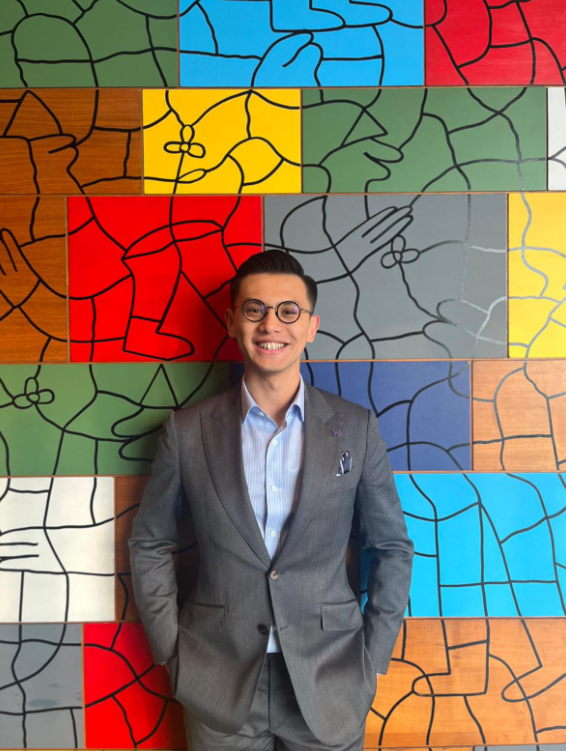A person wearing a gray suit, blue shirt, and round glasses stands smiling with hands in pockets. Behind them is a vibrant mural featuring abstract shapes and colorful sections in red, yellow, blue, and green.