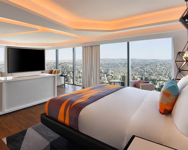 Modern hotel room with a large bed adorned with colorful pillows and a throw blanket, facing a flat-screen TV on a console. The room features floor-to-ceiling windows with cityscape views, wood flooring, and contemporary lighting on a unique ceiling design.