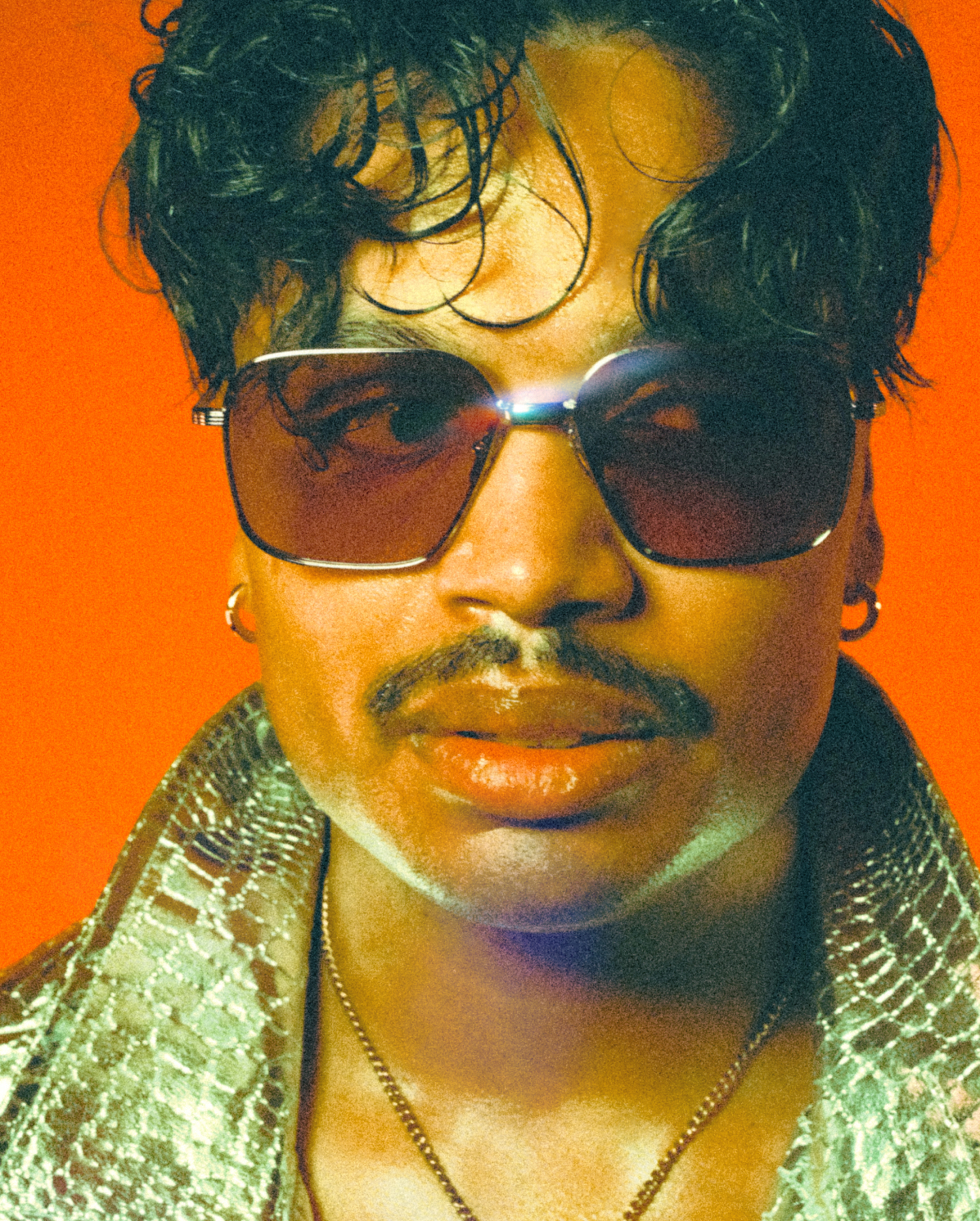 A person with curly hair and a mustache is wearing large sunglasses and a snakeskin-patterned jacket. They have a serious expression and are set against a bright red background.