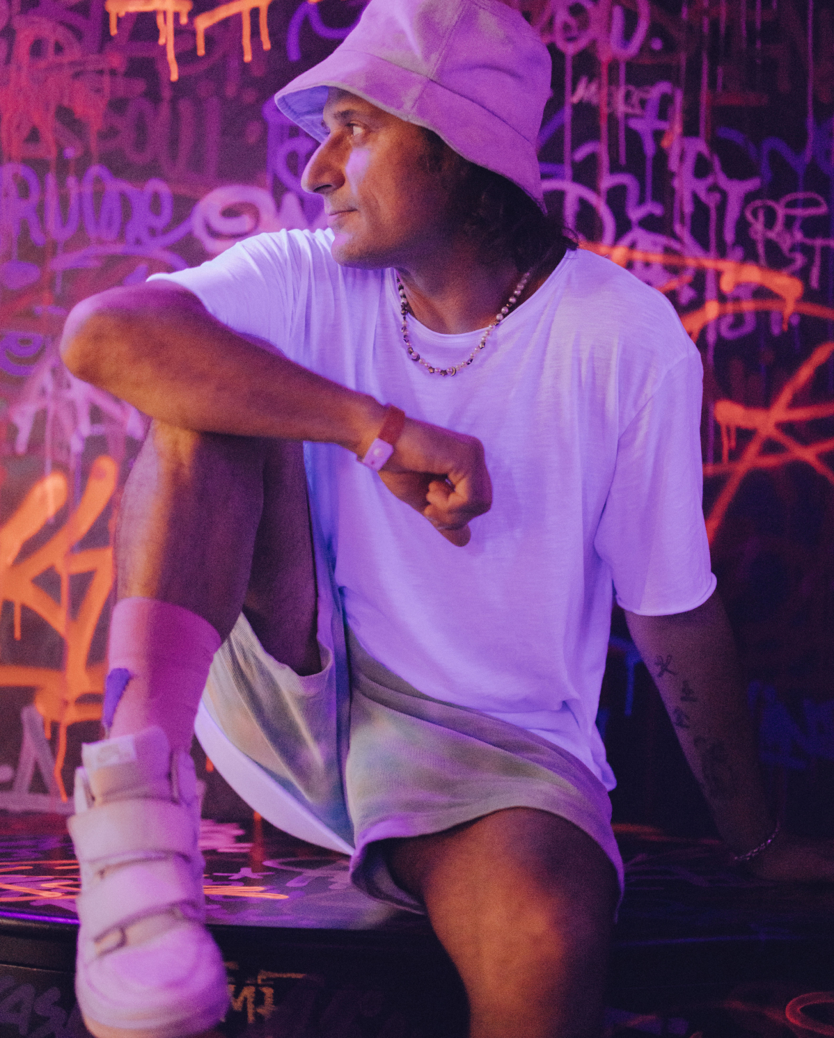A person wearing a lavender bucket hat, white t-shirt, pastel shorts, and white chunky sneakers sits on a surface with one leg bent and the other hanging off. They are in a dimly lit room with vibrant graffiti-covered walls and neon lighting.