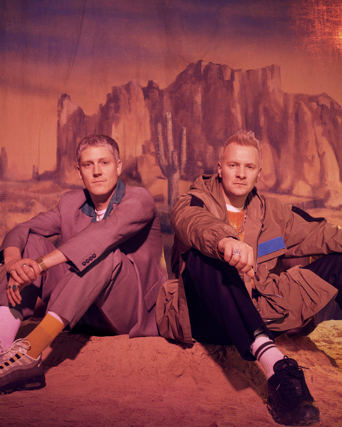 Two men sitting on sandy ground against a backdrop of a desert landscape with rocky formations. The man on the left wears a gray suit and sneakers, while the man on the right wears a brown jacket and black pants. Both have a relaxed and casual demeanor.