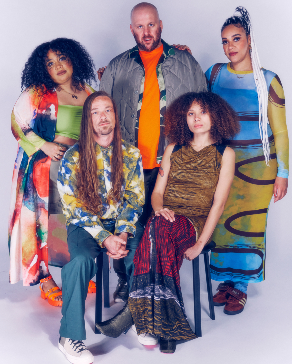 Five people pose together against a light background. One person is seated with others standing behind. Their outfits are colorful and varied, featuring patterns, bright colors, and unique styles. They display a mix of expressions, from smiles to calm and confident looks.