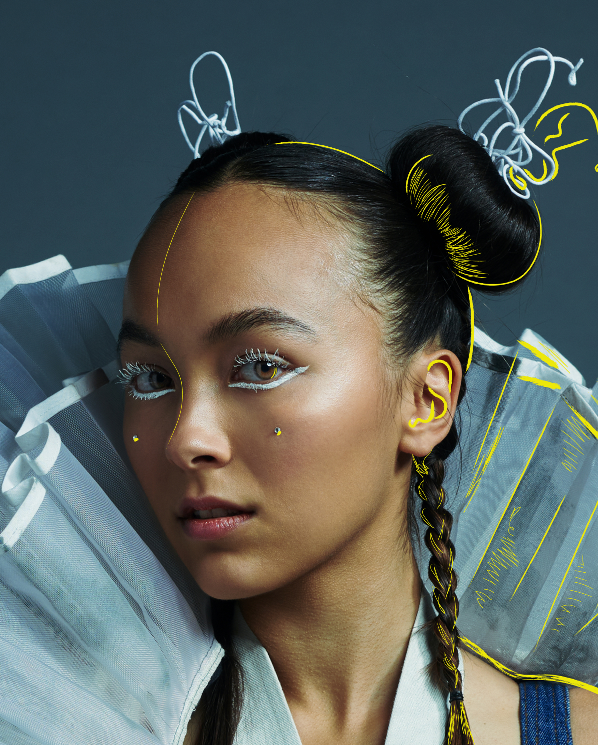 A portrait of a young person with intricate makeup and hair styling. Hair is styled in two braids and a bun with white and yellow accents. White makeup details highlight eyes and face. They wear a high-collared, ruffled garment against a gray background.