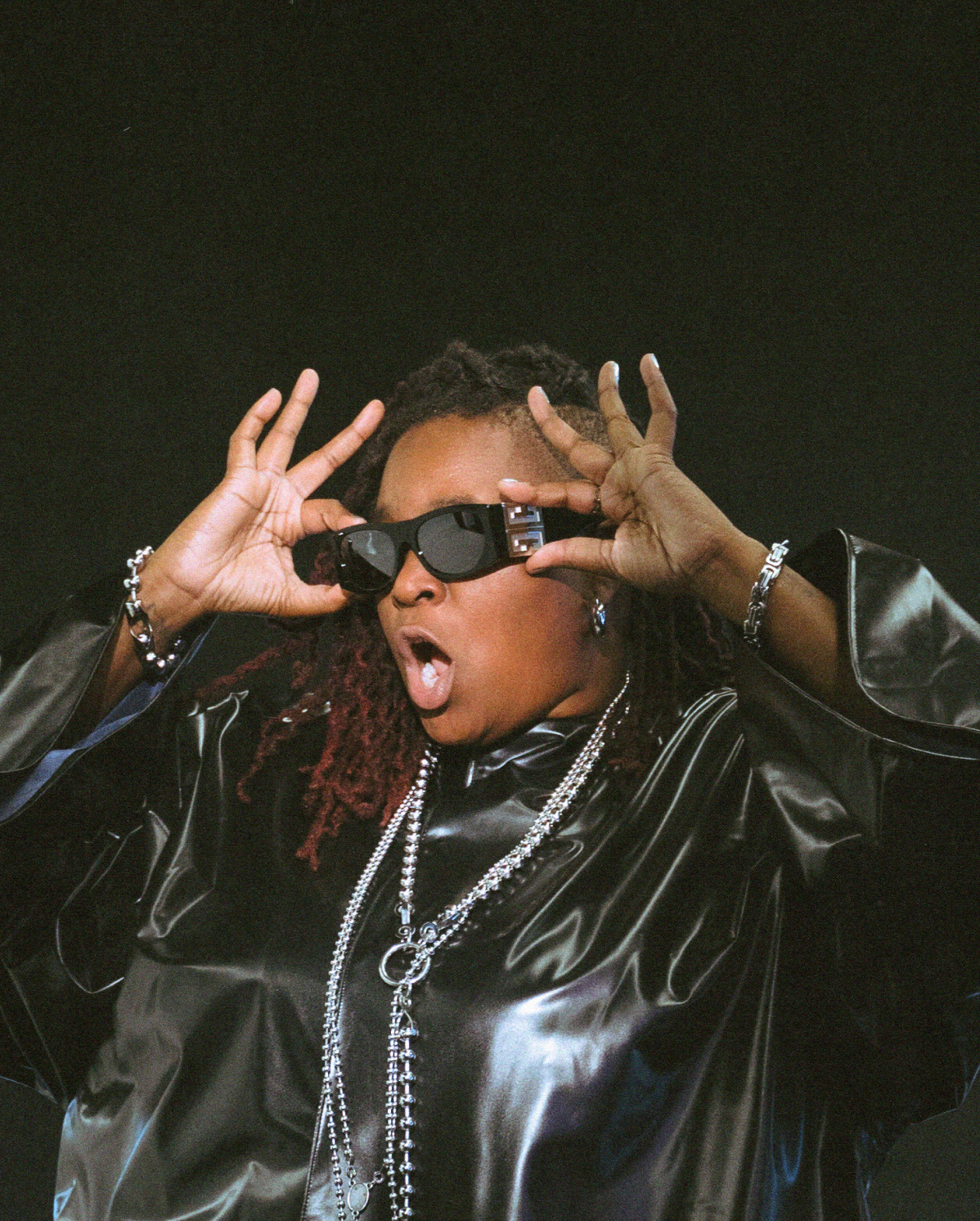 A person with long braids poses dramatically against a dark background, wearing black sunglasses and a shiny, oversized black jacket. They accessorize with multiple silver chains, bracelets, and rings, and their mouth is open in an expressive manner.