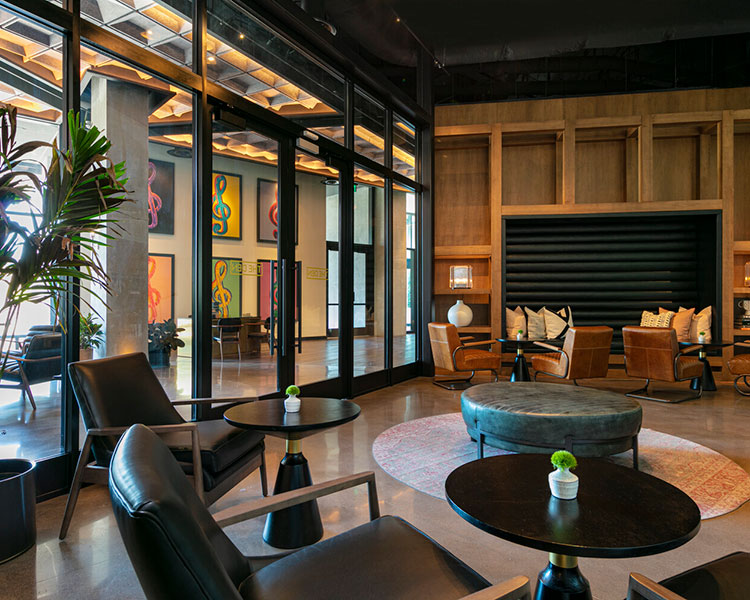 Modern lounge area with black leather chairs and round tables, with small potted plants as centerpieces. Wood and glass walls enclose the space, which features art on the walls, cushioned seating, and an overall cozy atmosphere. Large windows bring in natural light.