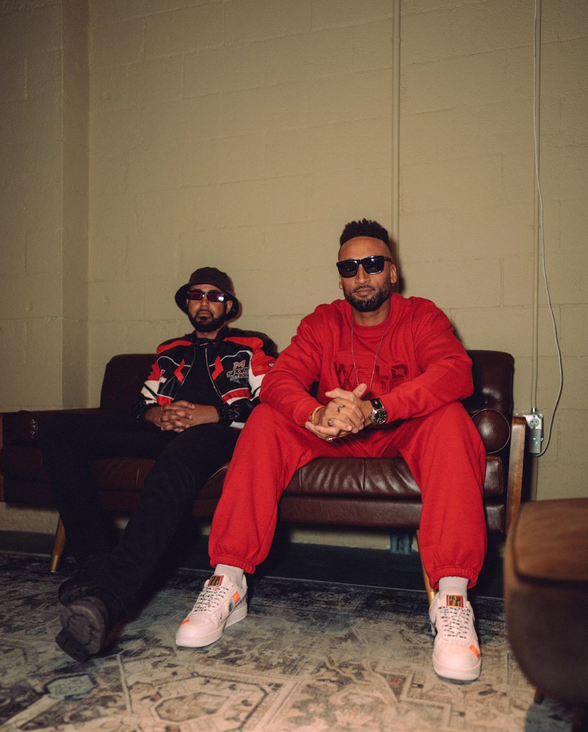 Two men sit on a brown leather couch in a dimly lit room. The man on the left wears a black bucket hat, sunglasses, a black and red jacket, and black pants. The man on the right wears sunglasses, a bright red tracksuit, and white sneakers. Both have a serious expression.