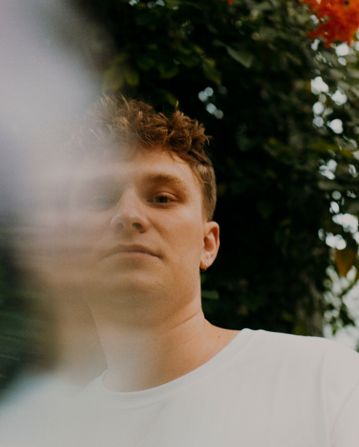 A person with short, curly hair is gazing slightly downward. They are wearing a white shirt, with a blurred section partially obscuring their face. The background features green foliage and a hint of colorful flowers. The overall tone is soft and slightly mysterious.
