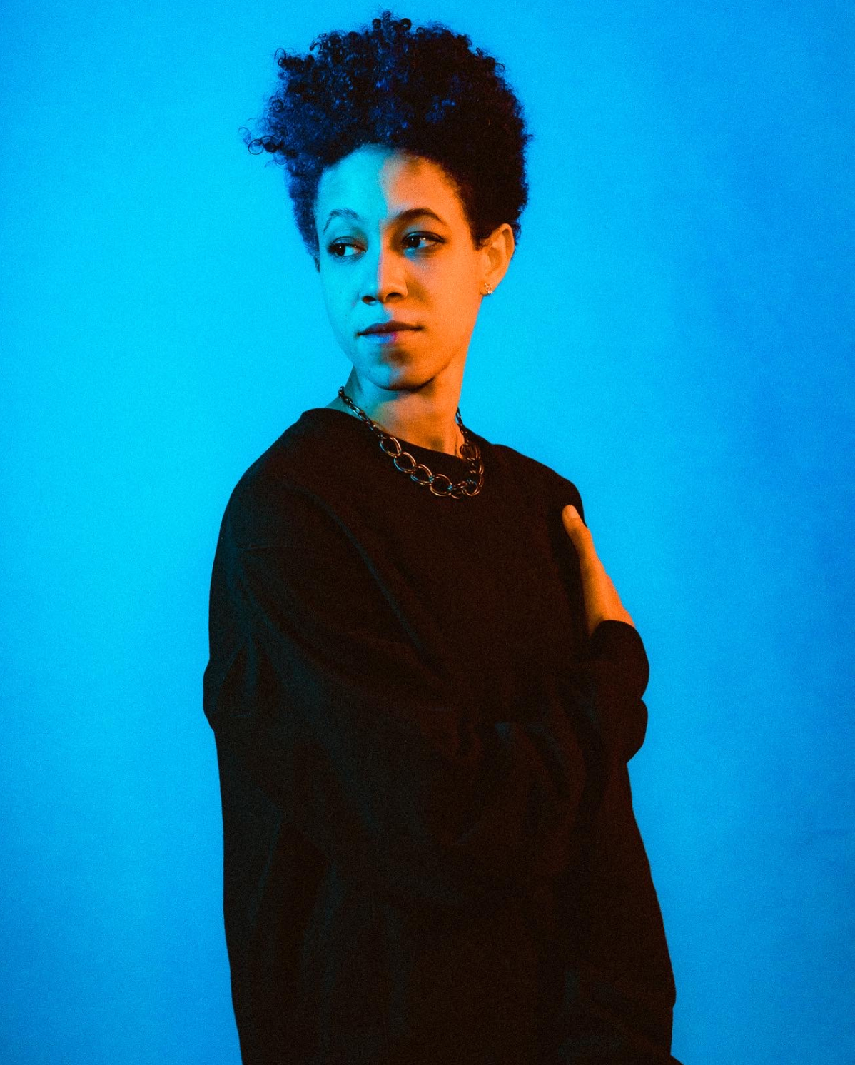 A person with short, curly hair stands against a blue background, wearing a dark sweater and a chain necklace. They have one arm bent, touching their opposite shoulder, while gazing slightly to the side with a contemplative expression.