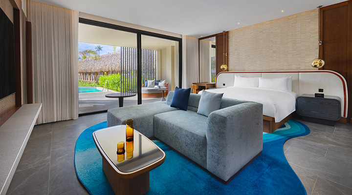Spacious hotel room with a large bed, modern gray sofa, coffee table with bottles, floor-to-ceiling windows, and a view of a patio, pool, and thatched roofs outside.