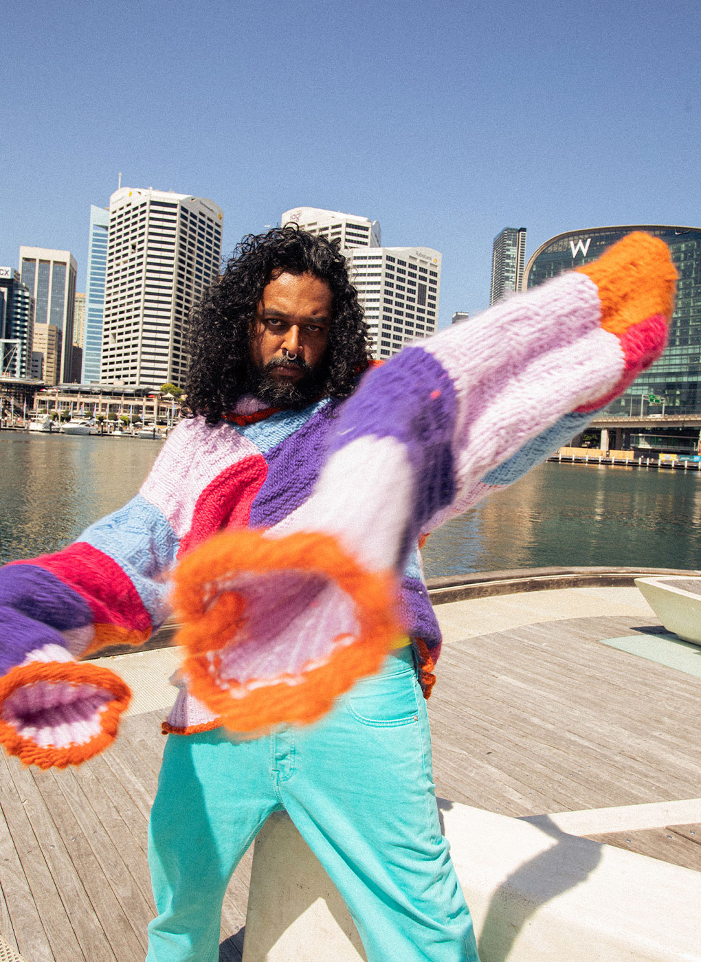 A man stands on a wooden dock near a body of water with modern buildings in the background. He has long, curly hair and is wearing a colorful, oversized sweater with flared sleeves and light blue pants. His right arm is extended, and he has a serious expression.