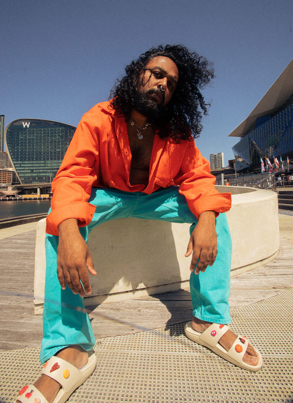 A person with long curly hair, wearing an unbuttoned orange shirt and blue pants, sits on a concrete structure by the waterfront on a sunny day. They are also wearing cream-colored sandals with colorful decorations. Modern buildings and a blue sky are in the background.