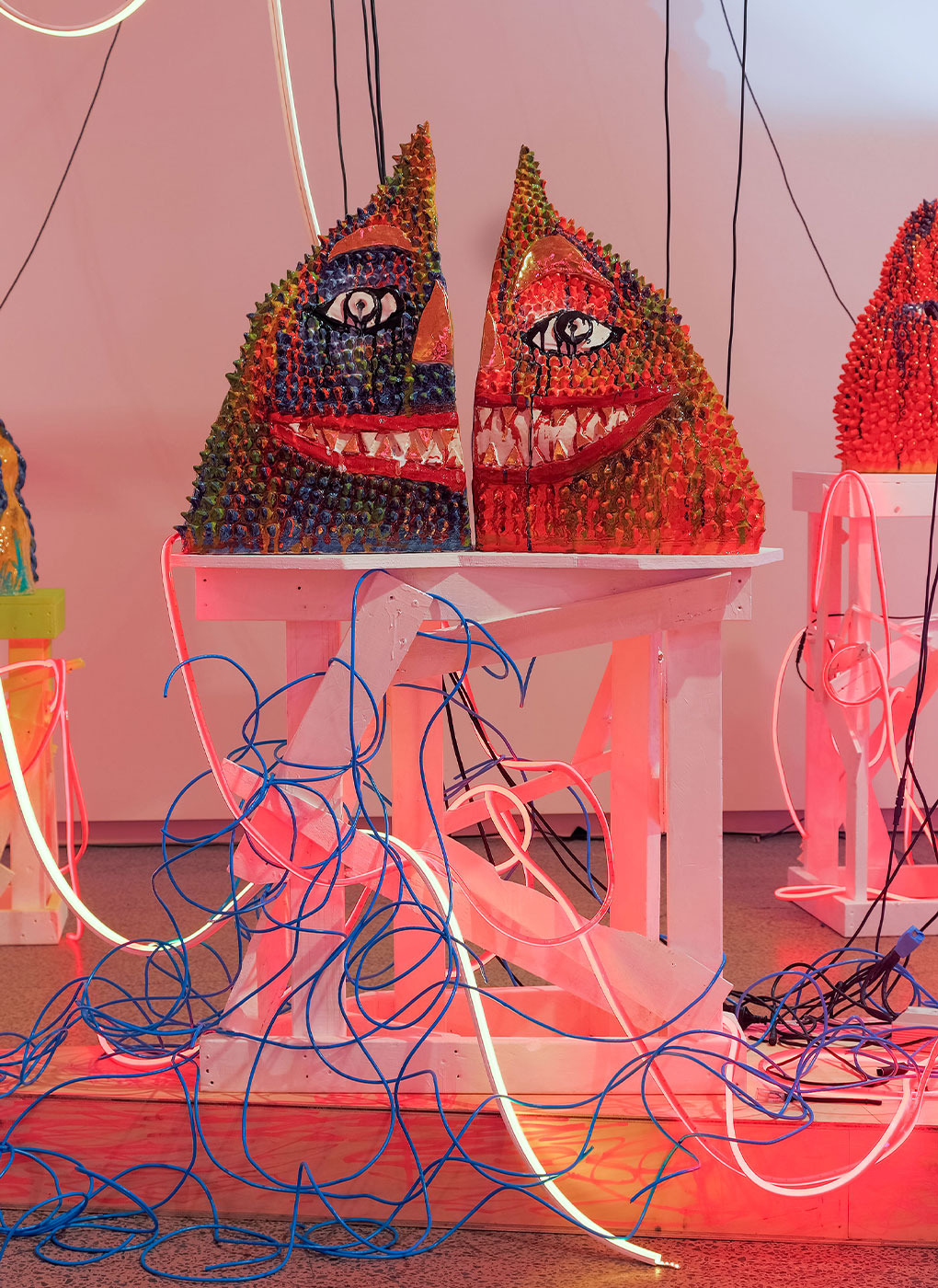 Colorful art installation features two face-like sculptures with intricate patterns and eye motifs. Positioned on a white table, the sculptures are surrounded by a chaotic array of blue and red wires, creating a vibrant, dynamic scene with a mix of textures and colors.