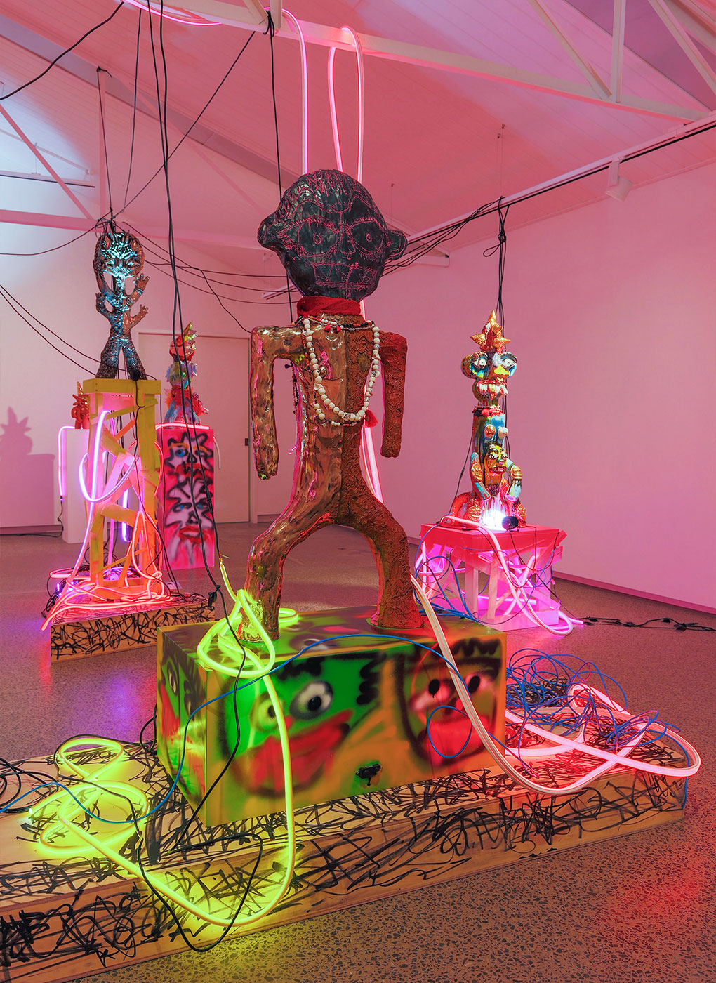 An art exhibit features three humanoid sculptures adorned with colorful neon lights and intricate patterns. The central figure stands prominently on a pedestal with outstretched arms, surrounded by electrifying cables and neon hues, set in a modern gallery space.