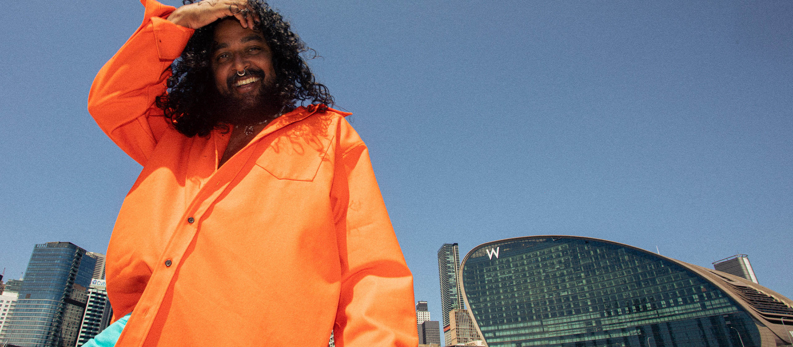 A person with long curly hair and a beard is wearing an orange jacket and standing outdoors. They are smiling and holding their left hand on their head. Modern buildings and a clear blue sky are visible in the background.