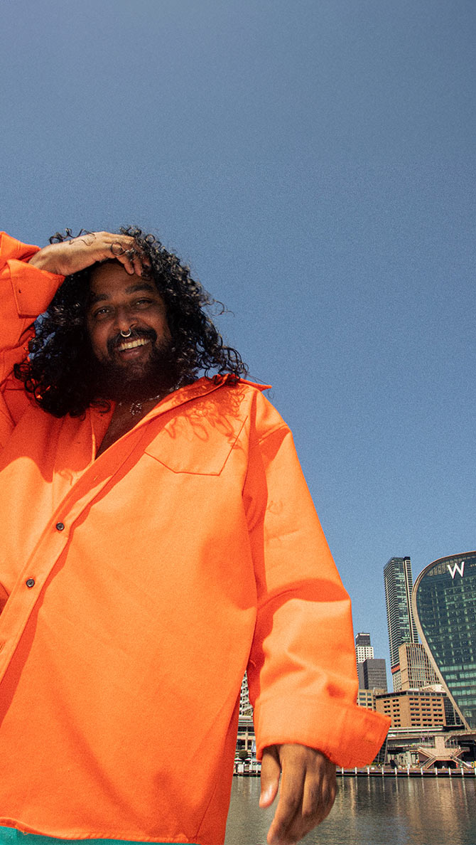 A person with long, curly hair and a beard smiles while wearing an orange shirt, standing outdoors with a city skyline and a clear blue sky in the background. They have one hand resting on their head.