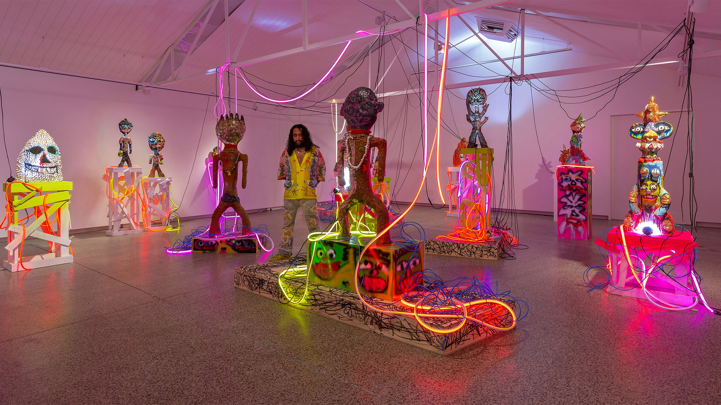 A person stands in a brightly lit art gallery featuring colorful, illuminated sculptures adorned with neon lights and wires. The sculptures are various abstract humanoid forms positioned on vibrant pedestals, creating a dynamic and visually stimulating environment.