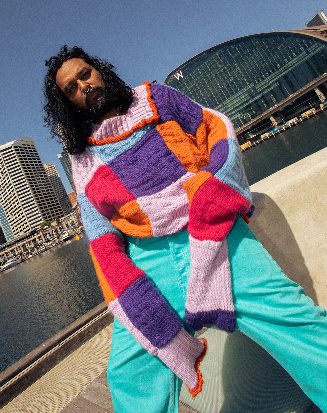 A person with long curly hair and a beard is wearing a colorful, oversized knit sweater and teal pants. They are posing outdoors near water, with a modern building featuring large glass windows in the background. The sky is clear and sunny.