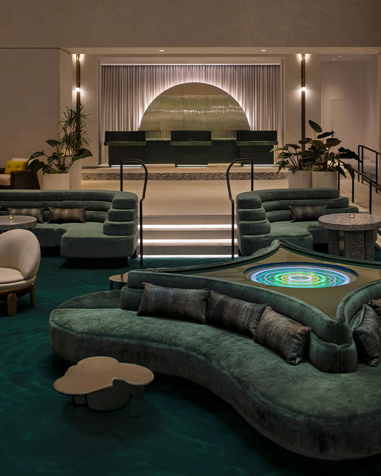 A modern lobby with a sunken seating area featuring plush, curvy green sofas and a multicolored circular light feature on a central table. Stairs with illuminated steps lead to a reception area with a large metallic art piece. Various plants decorate the space.