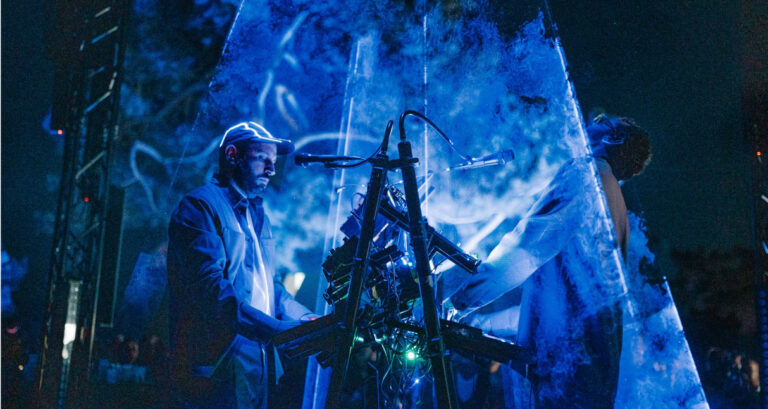 Two musicians perform on stage with electronic instruments and equipment. They are surrounded by a vibrant, blue-hued cloud of smoke. The scene is dimly lit, creating an atmospheric and immersive experience. One musician is wearing a cap and the other is looking up.