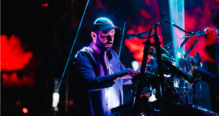 A musician stands behind a clear panel, wearing a cap and a jacket, focused on various electronic musical equipment. The background features red and black lighting, creating a dramatic ambiance. The scene suggests a live performance or studio session.