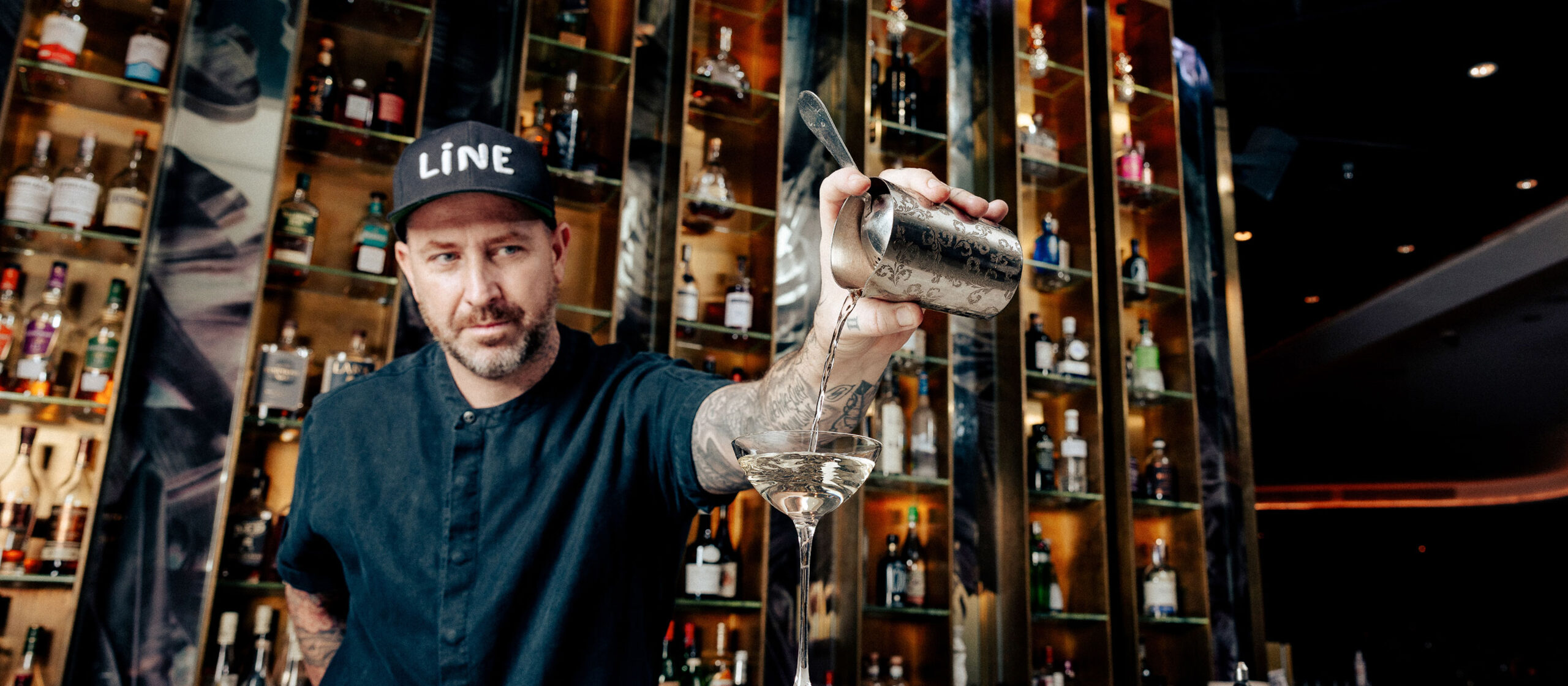 A bartender in a black shirt and baseball cap poured a cocktail from a shaker into a martini glass. Shelves filled with various liquor bottles are in the background, creating a lively and vibrant bar atmosphere.