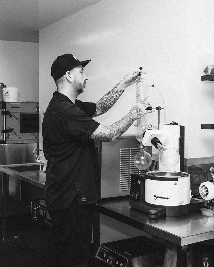 A person in a lab coat and cap is working with laboratory equipment. They appear to be adjusting a rotary evaporator in a clean, professional lab setting. Shelves with additional lab gear are visible in the background.