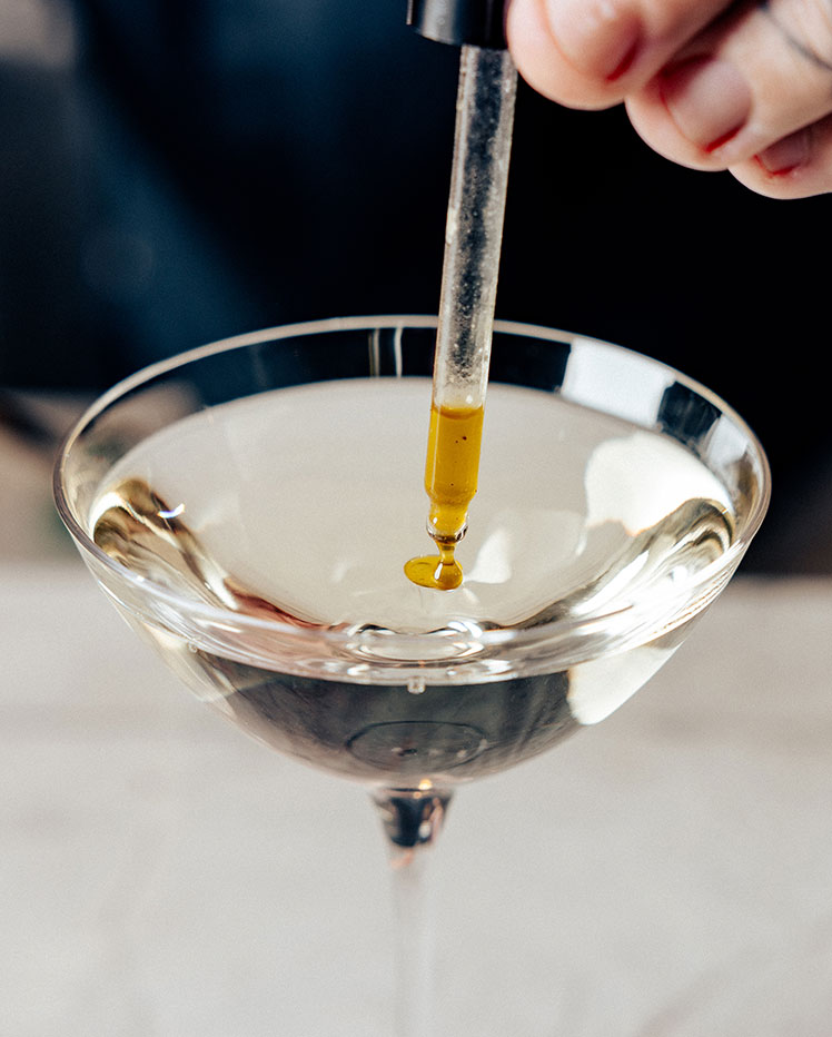 Close-up of a pipette dropping a yellow liquid into a clear glass of a transparent drink, possibly a cocktail or water. The background is blurred, focusing attention on the glass and pipette.